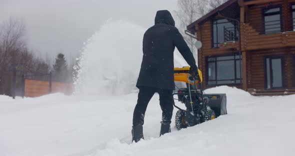 Man Cleans Snow With Snowplough at Wooden House Backyard in Winter