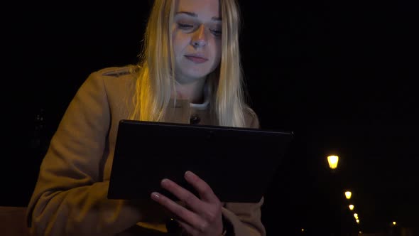 A Young Beautiful Woman Sits on a Bench and Works on a Tablet in an Urban Area at Night