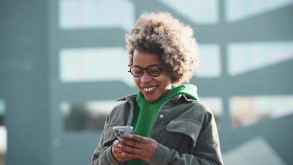 Smiling adult African woman wearing casual clothes texting by phone and looking to the side