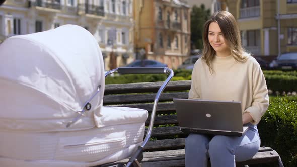 Happy Modern Mom Working Laptop Looking at Baby in Carriage Sitting Park Bench