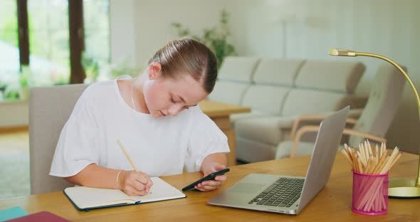 Teen Girl at the Desk in Front of Laptop Holds Smartphone and Writes Down Something Into Notebook alt