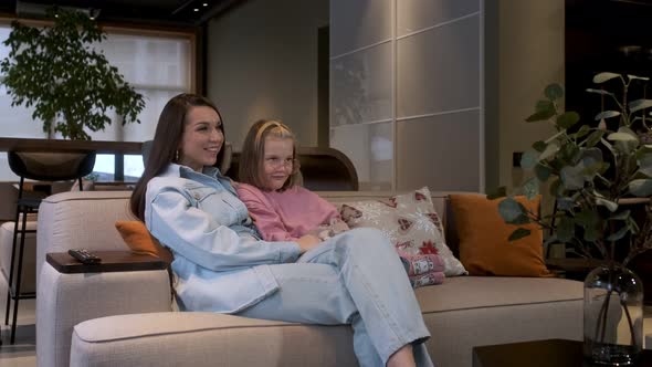 Happy young woman is sitting on cozy sofa with adorable little cute daughter. They watch TV