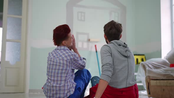 Back View Wide Shot of Relaxed Positive Couple Sitting on Floor Admiring House Painting on Wall