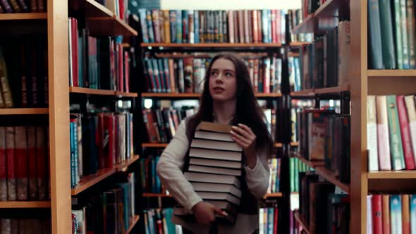 Student Girl Walks Through Library with Stack of Books in Hands Shelves with Book Front View