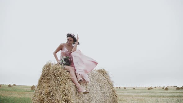 Attractive Happy Lady Sits in Pose on Large Haystack in Field Wind Blows Dress