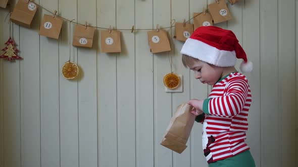 Boy in Pajamas Santa Cap Eating Sweet Candy on Paper Advent Calendar with Presents Background