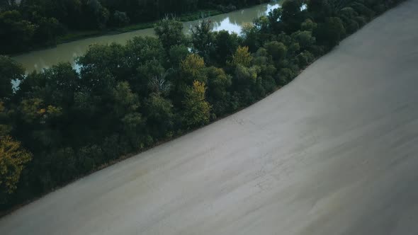 Drone shot over slow muddy river between fields and trees