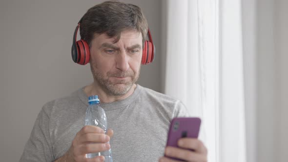 Positive Middle Aged Man in Headphones Drinking Refreshing Water and Listening to Playlist in