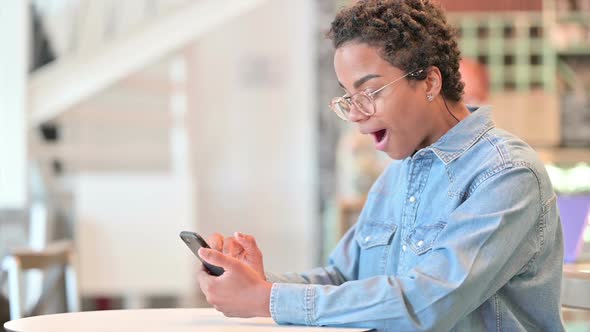 African Woman Celebrating Success on Smartphone at Cafe