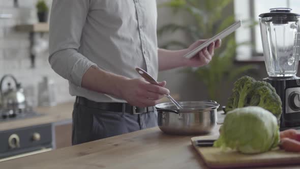 Unrecognizable Good-looking Man in the Shirt Cooking Soup Checking Recipe on the Tablet in the