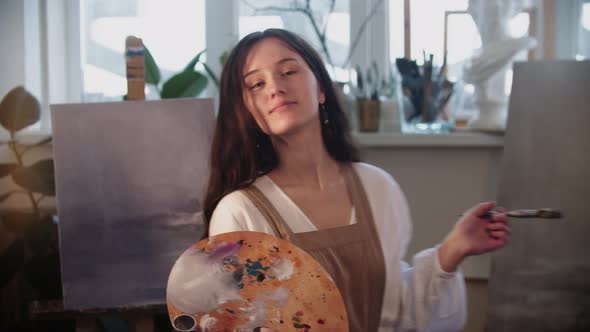 Young Smiling Woman Artist Walks in the Frame Holding Palette and Brush