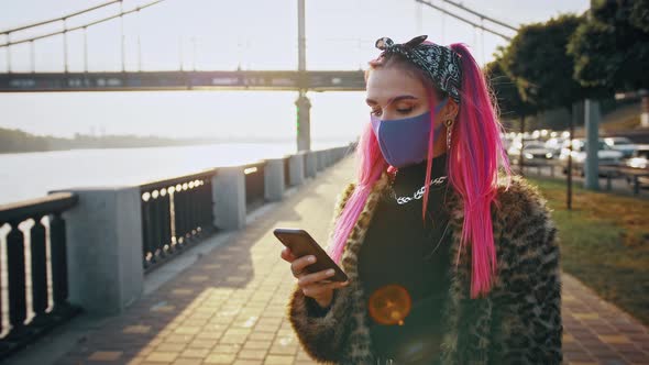 Young Hipster Lady in Informal Outfit and Protective Mask is Using Smartphone While Standing at