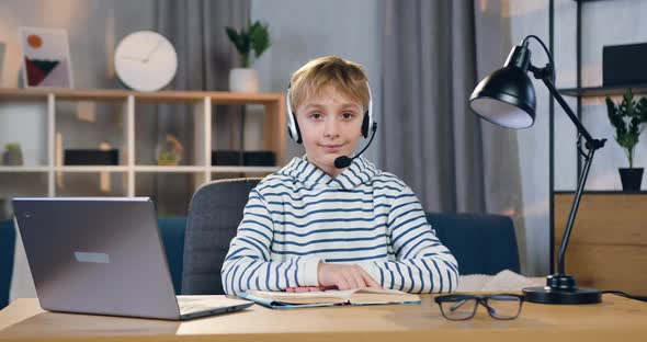 Smart Schoolboy in Headset which Sitting at his Workplace During Online Lesson and Looking at Camera