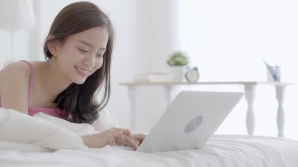 Young asian woman smiling lying on bed using laptop computer at bedroom for leisure.