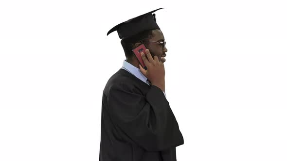 Smiling African American Male Student in Graduation Robe Talking on the Phone While Walking on White