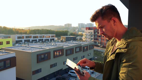 A Young, Handsome Man Works on a Tablet on a Balcony, Buildings Below