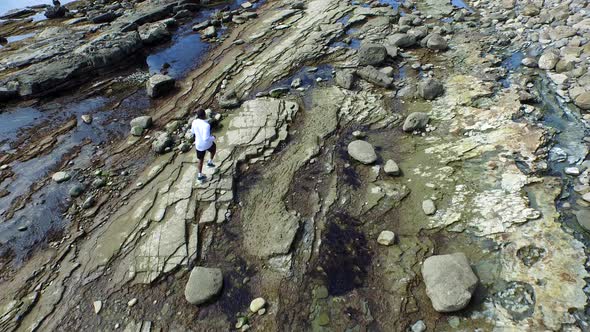 Aerial birds eye view shot of a young man running on a rocky ocean beach shoreline.