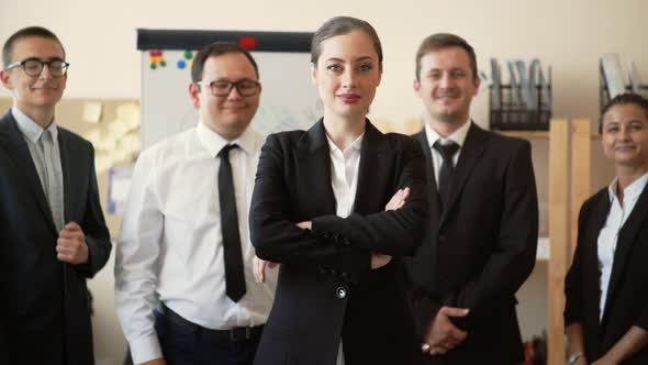 A Businesswoman Stands in the Office with Her Arms Crossed, Her Employees Stand Behind Her and Smile