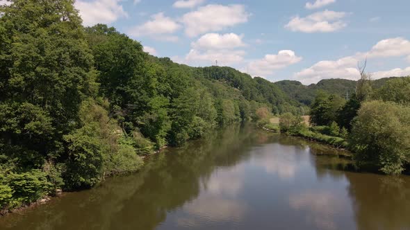 Calm, brown river surrounded with lush and leafy riverbanks underneath a blue sky with cumulus cloud