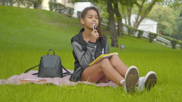 Pretty African American Schoolgirl Doing Homework on Sunny Green Lawn in Park. Portrait of