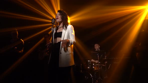 A Stylish Girl Vocalist Sings on Stage in a Vintage Microphone. In the Background Musicians Play