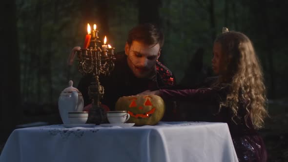 Portrait of Scary Vampire Opening Jackolantern for Little Girl Warming Hands in Forest Outdoors at