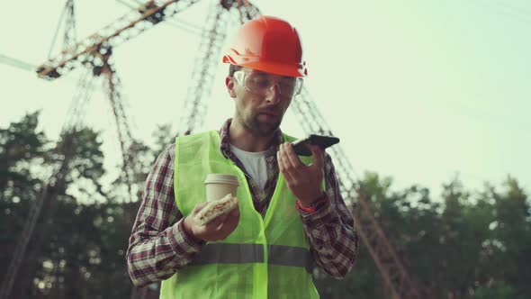 Male Worker Chief Electrician in Hard Hat Tries to Eat Sandwich and Drink Coffee but Needs to Work