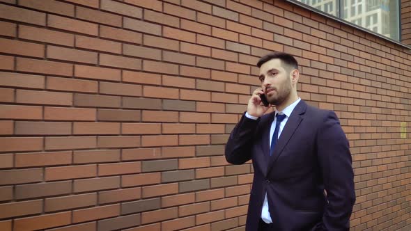 Businessman Dressed in Shirt Talking Smartphone While Walks on the Background of a Modern Brick Wall