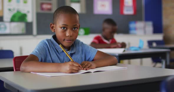 Portrait of happy african american schoolboy sitting at classroom, making notes, looking at camera