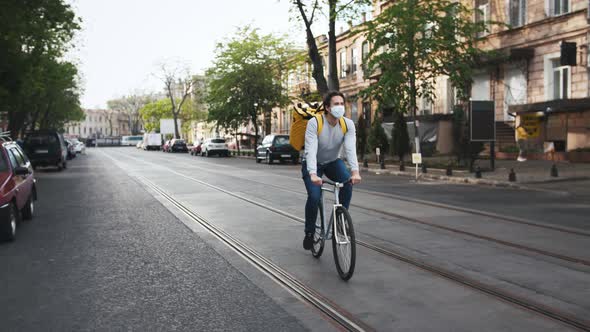 Courier with Yellow Backpack and Protective Mask Rides a Bicycle on the Street Through the City with