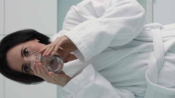 Vertical video of thirsty smiling 35s woman in white bathrobe drinking water