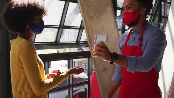 Mixed race male cafe worker wearing face mask taking temperature of female coworker