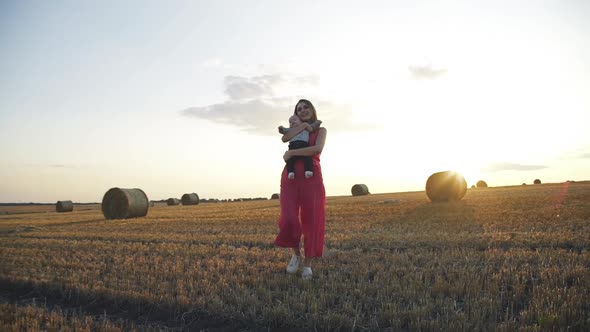 Happy Mother with Cute Baby Son in Hands Walking on Spacious Hay Field at Sunset