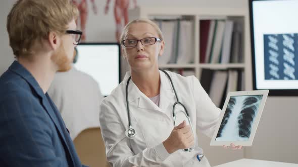 Doctor Explaining X-ray on Digital Tablet To Patient at Desk in Hospital