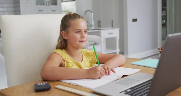 Happy caucasian girl at home, sitting at desk writing during online school lesson using laptop