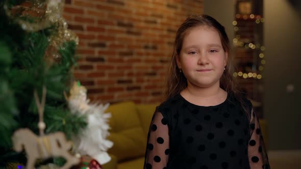 Child portrait looking at camera near christmas tree at home indoors. Young cute female girl