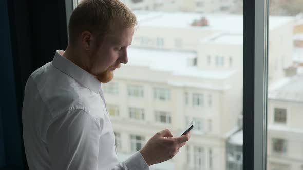 businessman uses smartphone while standing by the window overlooking buildings