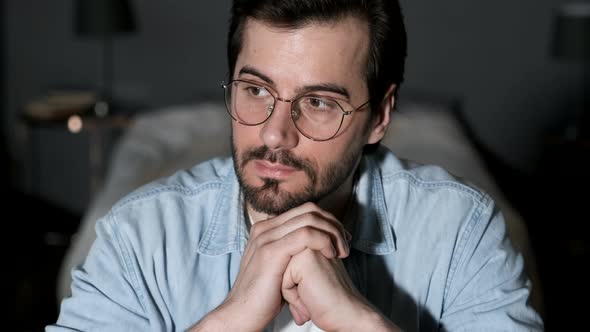 A young Man Thinks Looks Away, Uses a Laptop Works Online in a Cozy Dark Room at home in the Evening