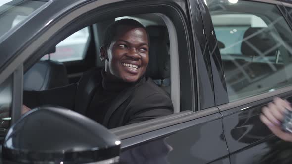 Portrait of Happy African American Man Sitting in Vehicle Salon and Taking Keys From Caucasian Car
