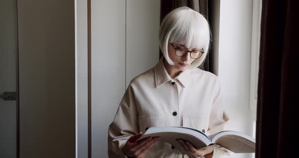 Beautiful Woman with Glasses Holds a Book in Her Hand and Looks Out the Window