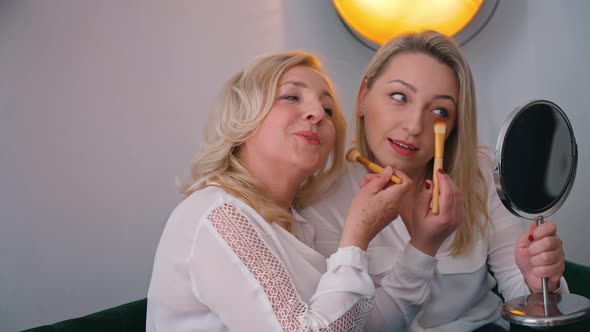 Two Blondehaired Focused Attractive Women Fixing Their Makeup with Gib Fluffy Brushes Looking at a