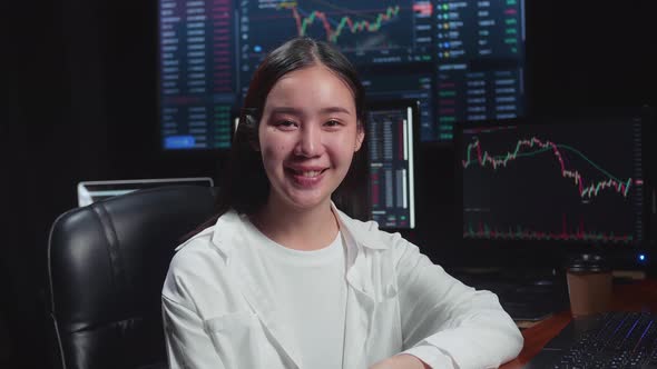 Asian Female Stock Market Broker Working On Computer And Smiling To Camera With Analysing Graphs