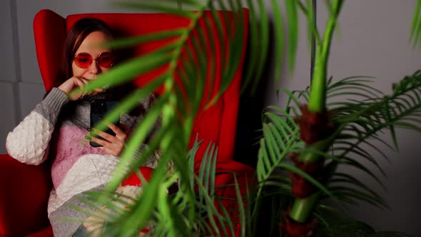 Woman in Stylish Sunglasses Sitting in Armchair with Smartphone