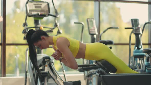 Young Hispanic Woman in a Yellow Tracksuit Performs a Hyperextension Exercise to Train the Muscles