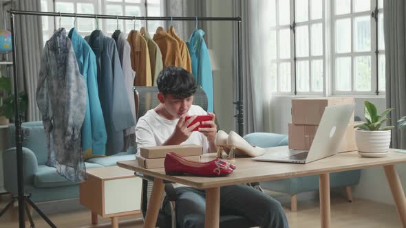 Asian Man Online Seller Taking Photos With Mobile Phone While Using Computer For Selling Clothes