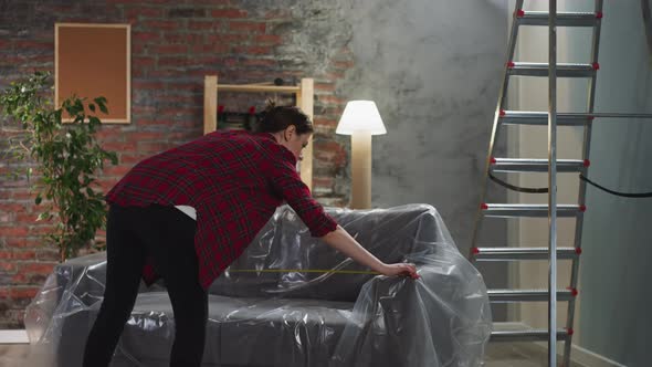 Woman Measures Sofa Length Covered with Foil in Living Room