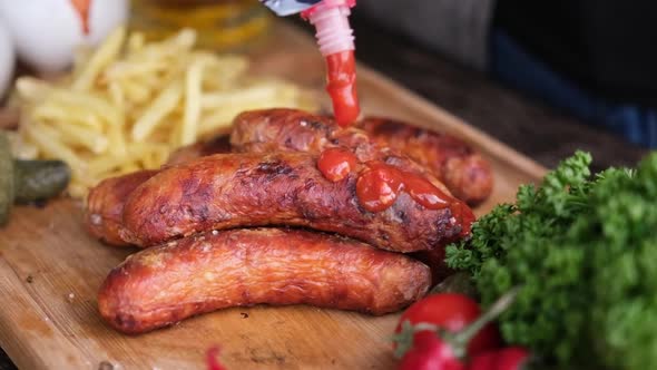 Pouring Tomato Sauce on Tasty Grilled Sausages on Wooden Serving Board with French Fries and Sauces