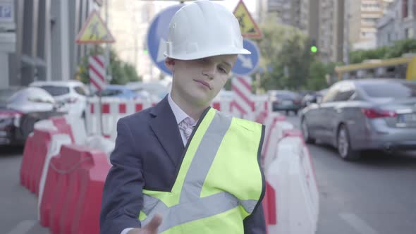 Tired Little Boy in Constructor Helmet on His Head, and Uniform Looking in Camera Showing Thumb Up