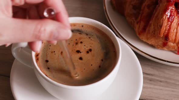 A Man Stirs Sugar in a Cup of Aromatic Coffee with Glass Spoon on Wood Table