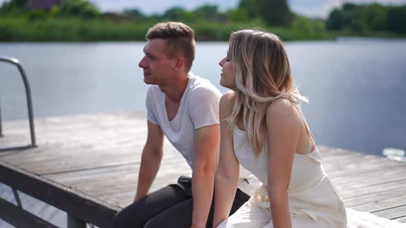 Young Romantic Caucasian Couple Sitting on Wooden Pier Talking Dating on Sunny Summer Day Outdoors